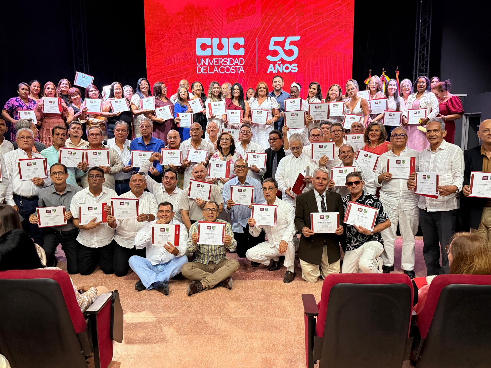 Grupo numeroso de personas posando sobre un escenario, cada una sosteniendo un certificado. Al fondo se ve una pantalla roja brillante con el logo y nombre de la "CUC Universidad de la Costa" y el texto "55 AÑOS"
