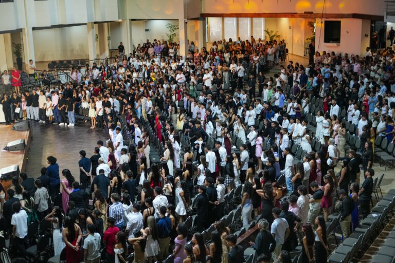 Auditorio lleno de estudiantes Los asistentes están vestidos con ropa semi-formal y formal, como vestidos, camisas y trajes, y se encuentran de pie entre filas de sillas ocupadas