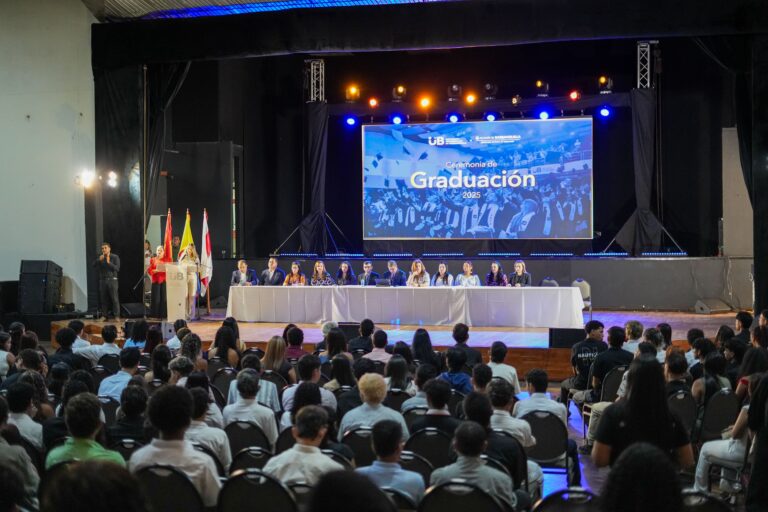 ceremonia de graduación en un auditorio. En el escenario, hay una mesa larga cubierta con mantel blanco, detrás de la cual están sentadas varias personas, posiblemente autoridades académicas. A la izquierda del escenario, una persona está hablando desde un podio que lleva el logo “UB”