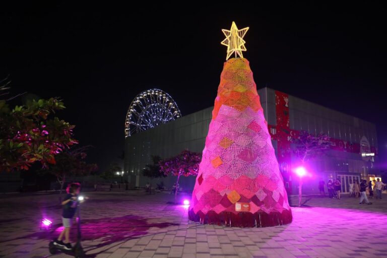 árbol navideño grande, ubicado en un espacio abierto durante la noche. El árbol tiene forma cónica y está completamente cubierto por piezas tejidas en diferentes tonos de rosa, rojo, fucsia y naranja, formando un diseño artesanal. En la parte superior hay una estrella luminosa dorada.