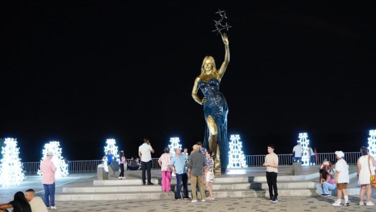 Estatua dorada y azul que representa a Sofia Vergara, ubicada en el Gran Malecón. Hay varias personas observando y tomando fotos, el fondo está decorado con figuras luminosas en forma de árboles navideños, dispuestas en filas simétricas, creando un ambiente festivo.