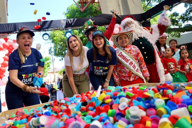 En primer plano hay una mesa grande llena de tapas plásticas de colores (rojas, azules, verdes, amarillas, entre otras), y varias personas están participando activamente, extendiendo las manos hacia las tapas. Algunas llevan camisetas con el texto visible “Barranquilla” y gorras alusivas a la ciudad y a la Navidad.