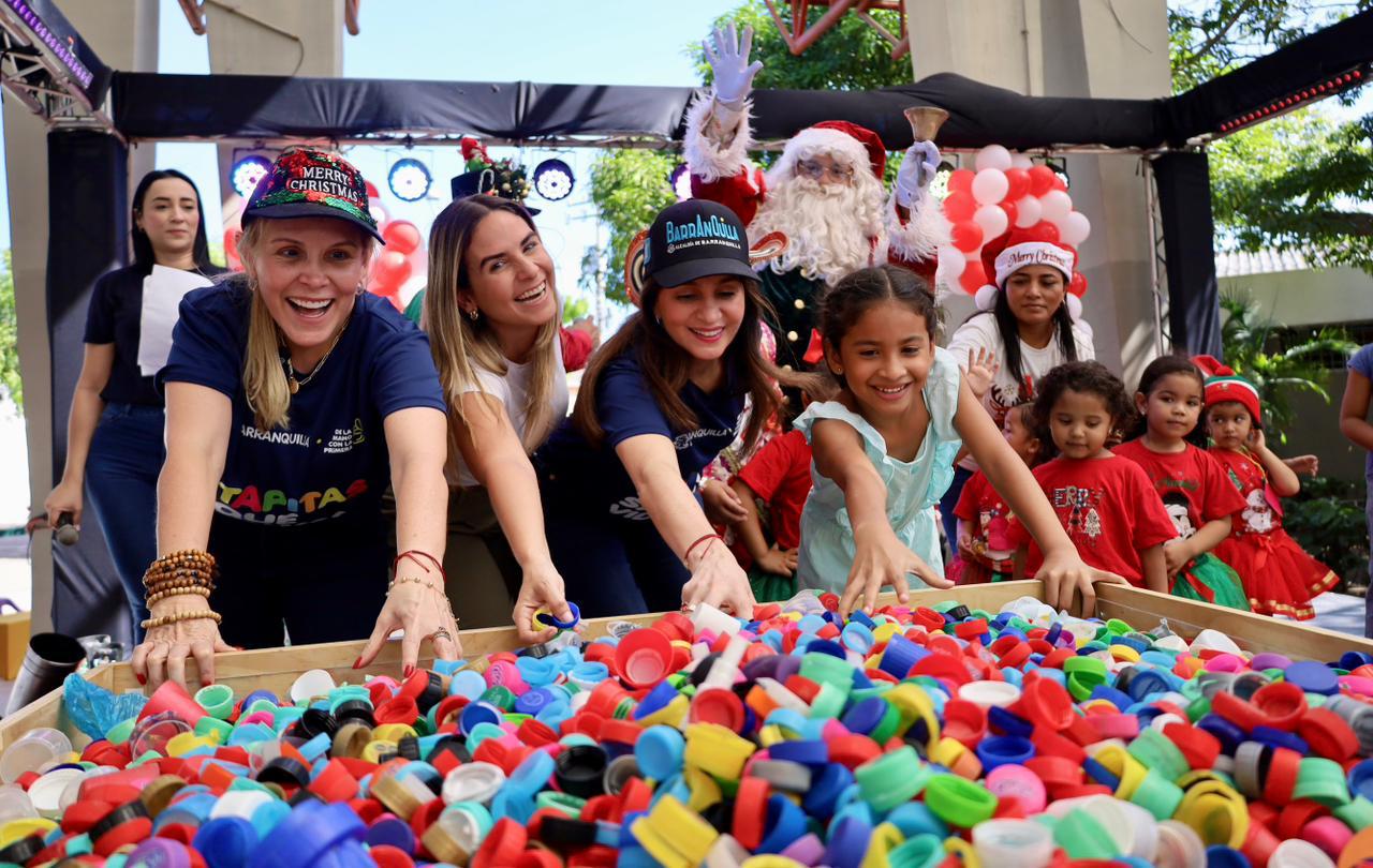 Actividad grupal en un espacio decorado con temática navideña. En primer plano hay una mesa grande llena de tapas plásticas de colores (rojas, azules, verdes, amarillas, entre otras), y varias personas están extendiendo las manos hacia ellas, participando activamente en la dinámica.