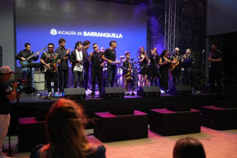 Escenario iluminado, donde un grupo numeroso de músicos y artistas se encuentra sobre una tarima realizando una presentación en vivo. En el fondo destaca una gran pantalla LED con el texto “ALCALDÍA DE BARRANQUILLA