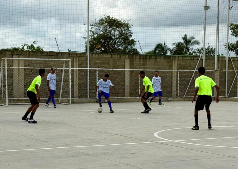 un grupo de jóvenes jugando un partido de fútbol sala en una cancha al aire libre. Dos equipos, diferenciados por sus camisetas uno con camisetas blancas y medias azules, y el otro con camisetas amarillas fluorescentes disputan la posesión del balón