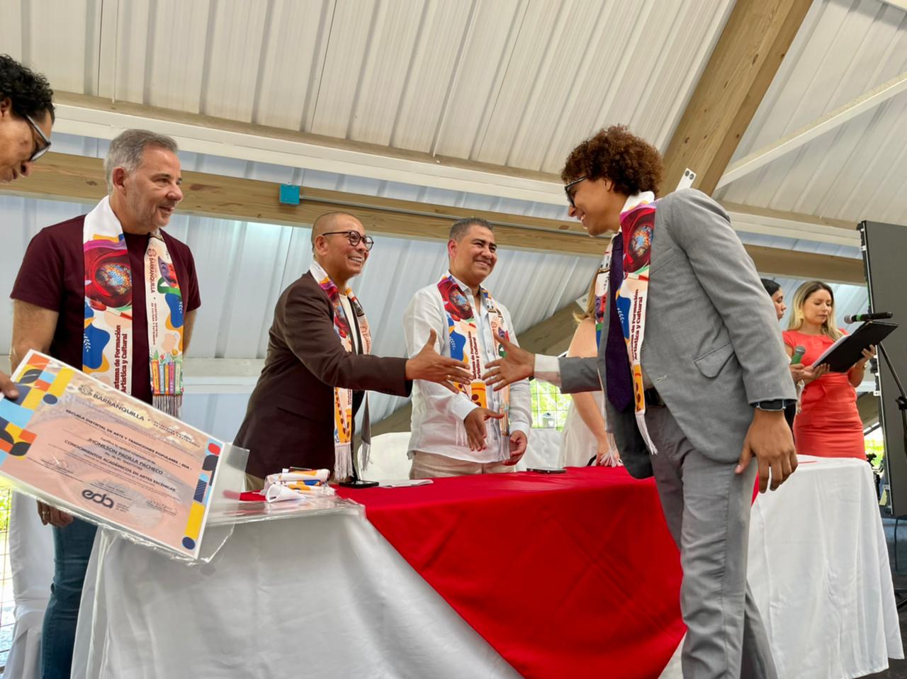 joven vestido con traje gris estrechando la mano de uno de los miembros de la mesa principal durante una ceremonia de graduación. Todos llevan estolas coloridas características de la EDA. Los miembros de la mesa, también vestidos de manera formal, sonríen mientras entregan certificados.