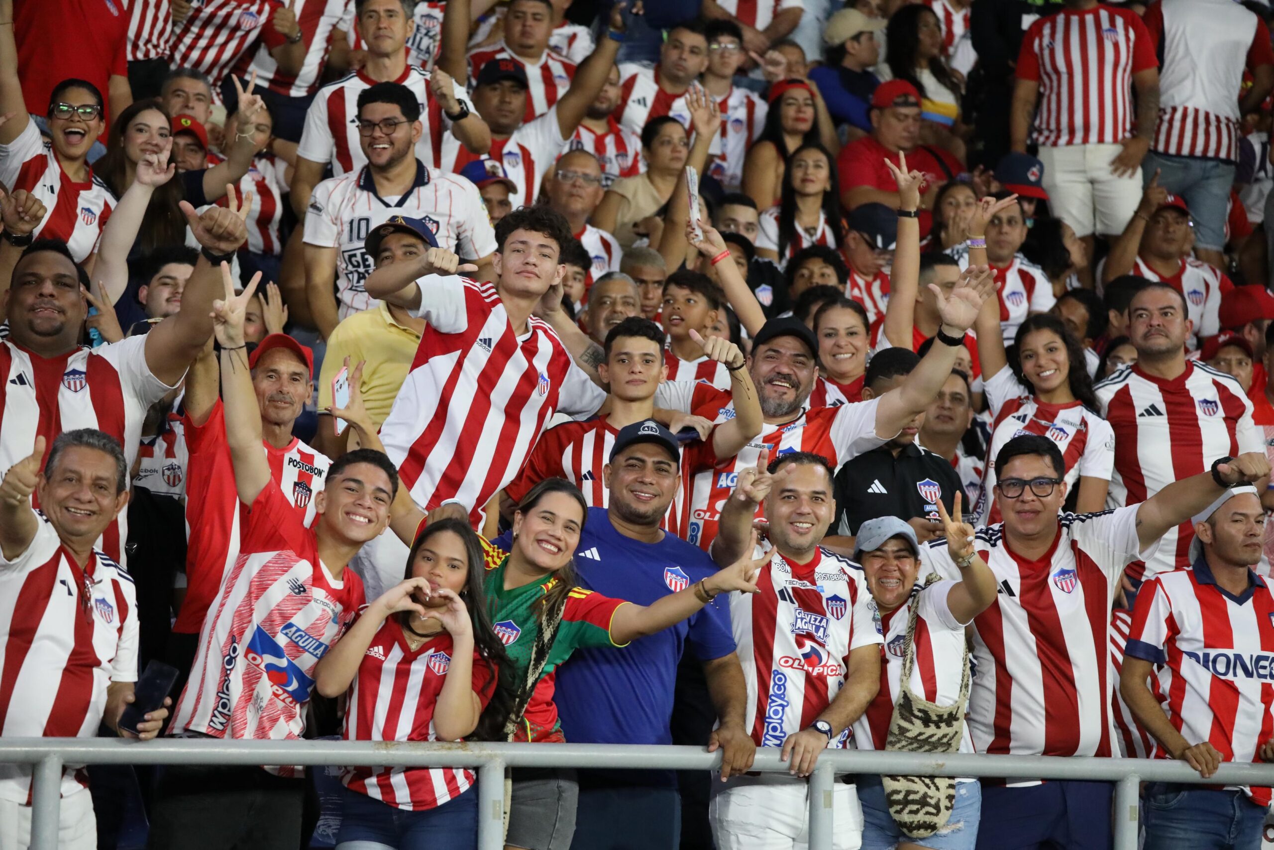 multitud de aficionados en las gradas de un estadio, todos vistiendo camisetas con los colores rojo y blanco, característicos del equipo Atlético Junior. Se observan varias personas levantando las manos, haciendo gestos de celebración y mostrando entusiasmo por el partido