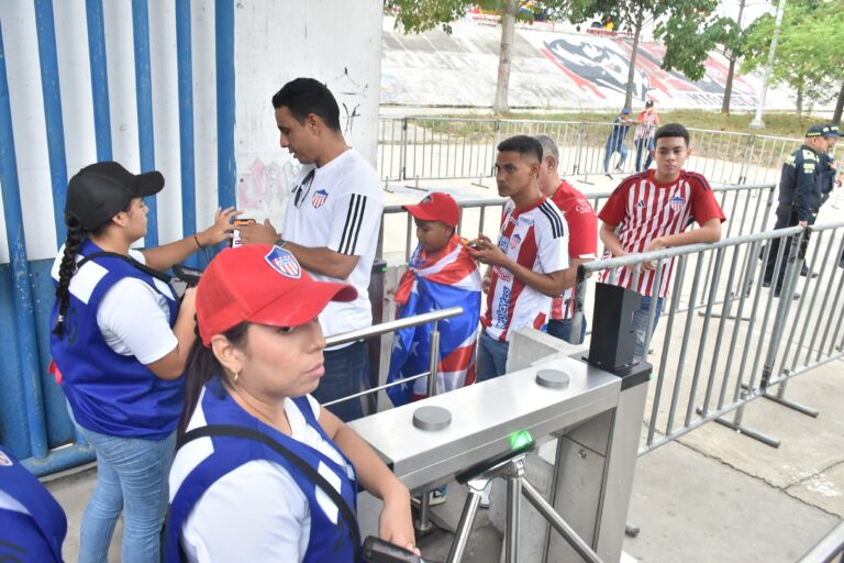 Varios aficionados del Junior ingresando al estadio Metropolitano a través de un torniquete de acceso. Un miembro del equipo de logística, verifica las entradas en el dispositivo móvil de un asistente.