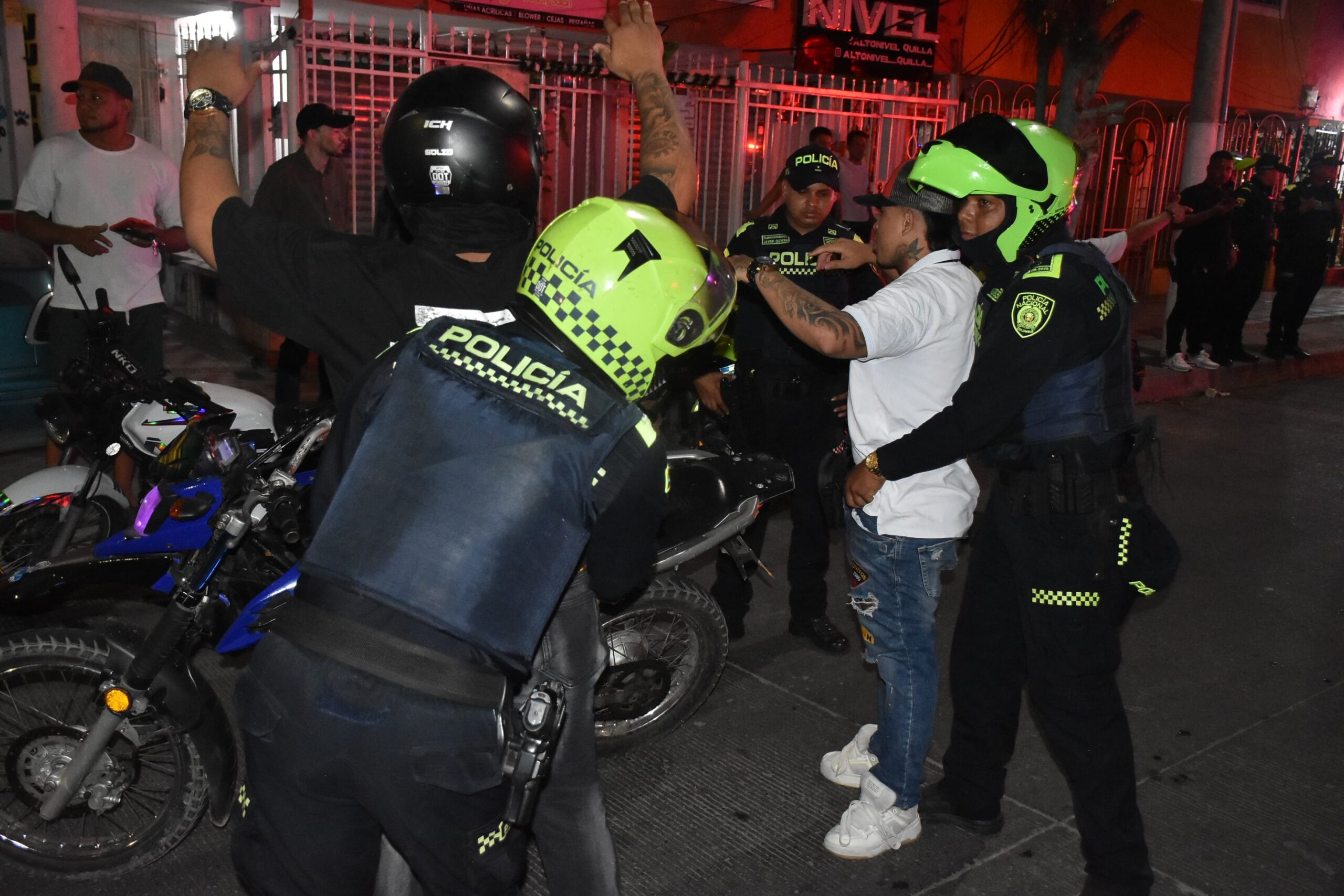 Los agentes están realizando requisa a dos personas. Una de ellas está de pie con los brazos levantados, mientras un policía lo revisa.