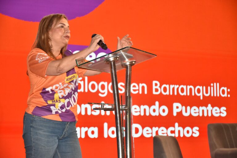 La Jefa de la Oficina Distrital de la Mujer, Equidad y Género, en un escenario frente a un atril transparente con micrófono. El fondo es una pantalla grande de color naranja con texto en blanco