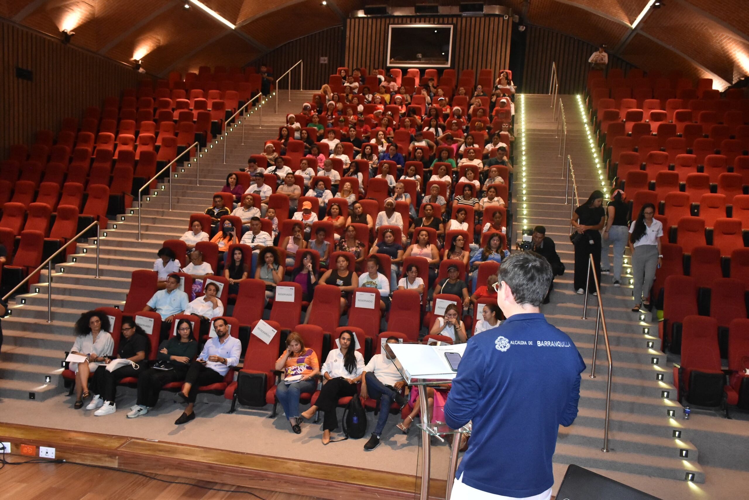 Auditorio con muchas sillas rojas dispuestas en filas ascendentes. En el escenario, hay una persona hablando desde un atril con documentos, dirigiéndose al público. La mayoría de las sillas del centro están ocupadas por personas que parecen estar escuchando la presentación.