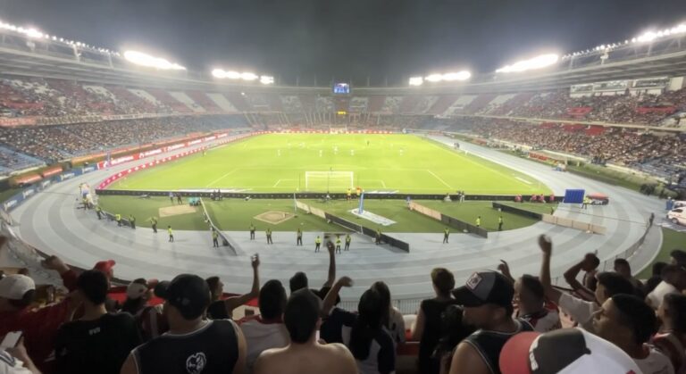 Vista panorámica de un estadio iluminado durante un partido de fútbol, con aficionados animando desde las gradas detrás de la portería.
