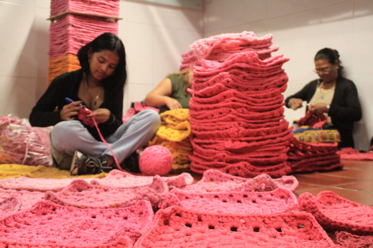 Mujeres sentadas en el suelo, tejiendo piezas de crochet con hilo grueso tipo trapillo en tonos rosados y amarillos.