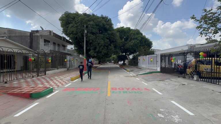 una cuadra totalmente embellecida del barrio Boyacá. La calle está limpia y organizada, con los bordes de los andenes pintados en colores rojo, verde y amarillo. A ambos lados se ven casas con rejas decoradas con globos en los mismos colores.