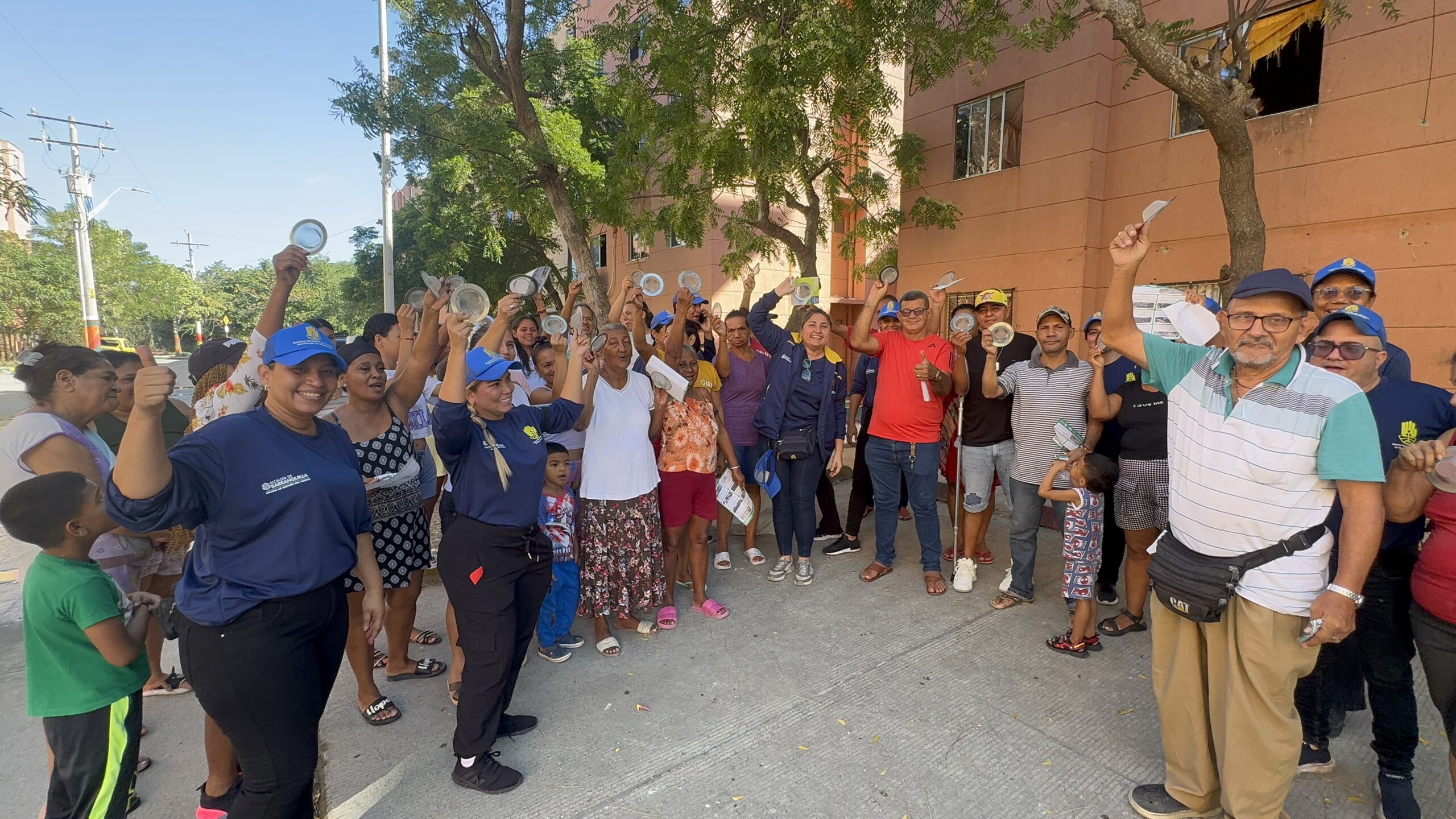 Personas reunidas en una calle peatonal, junto a un edificio, participan con entusiasmo levantando utensilios de comida, reflejando un ambiente alegre y de participación comunitaria.