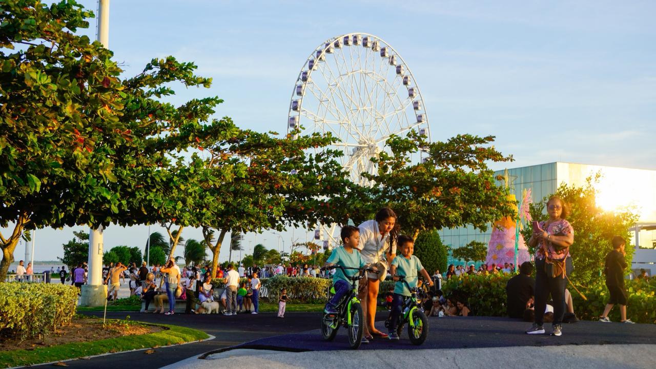 Parque lleno de gente disfrutando hay dos niños montados en bicicleta sosteniendo por una adulta en el fondo se ve una gran rueda de la fortuna, que destaca entre los arboles verdes.