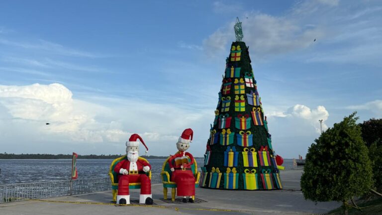 Un árbol de Navidad monumental decorado con figuras coloridas en forma de regalos se encuentra en el Malecón del Río, acompañado por dos figuras grandes de Papá Noel sentadas en sillones, todo bajo un cielo despejado y junto al río.