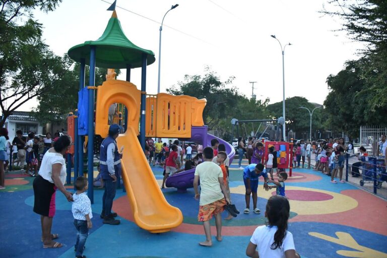 Parque infantil al aire libre con una gran cantidad de personas reunidas. En el centro se observa una estructura de juegos colorida, compuesta por un tobogán naranja, una escalera y un techo verde en forma de cono. También hay otros elementos como un tobogán morado y paneles interactivos para niños.