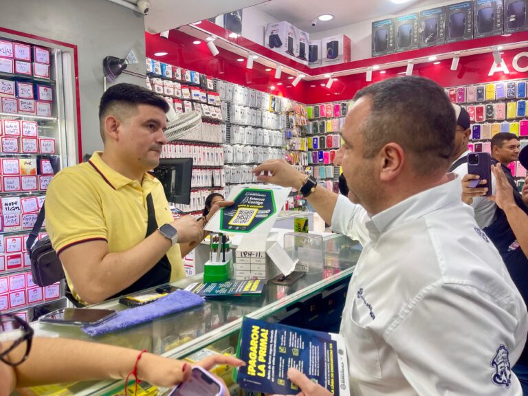 En primer plano, sobre un mostrador de vidrio, se observa material promocional con un código QR impreso en un cartel y un folleto Varias personas participan en la interacción, entregando y recibiendo la información