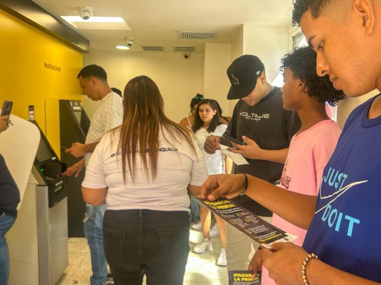 Cajero automático sobre un panel amarillo que dice “Multifuncional”. Varias personas están de pie formando fila frente al cajero, algunas usando dispositivos móviles y otras sosteniendo un volante informativo que incluye el texto “¡PAGAR LA PRIMA!”
