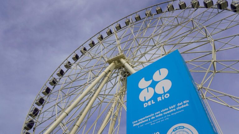 Una gran rueda de la fortuna metálica con cabinas distribuidas en su borde, vista desde un ángulo bajo contra un cielo despejado gris azulado. Hay un cartel azul con texto blanco que promociona la atracción, indicando que ofrece una experiencia con vista panorámica de 360° del Río Magdalena y la ciudad.