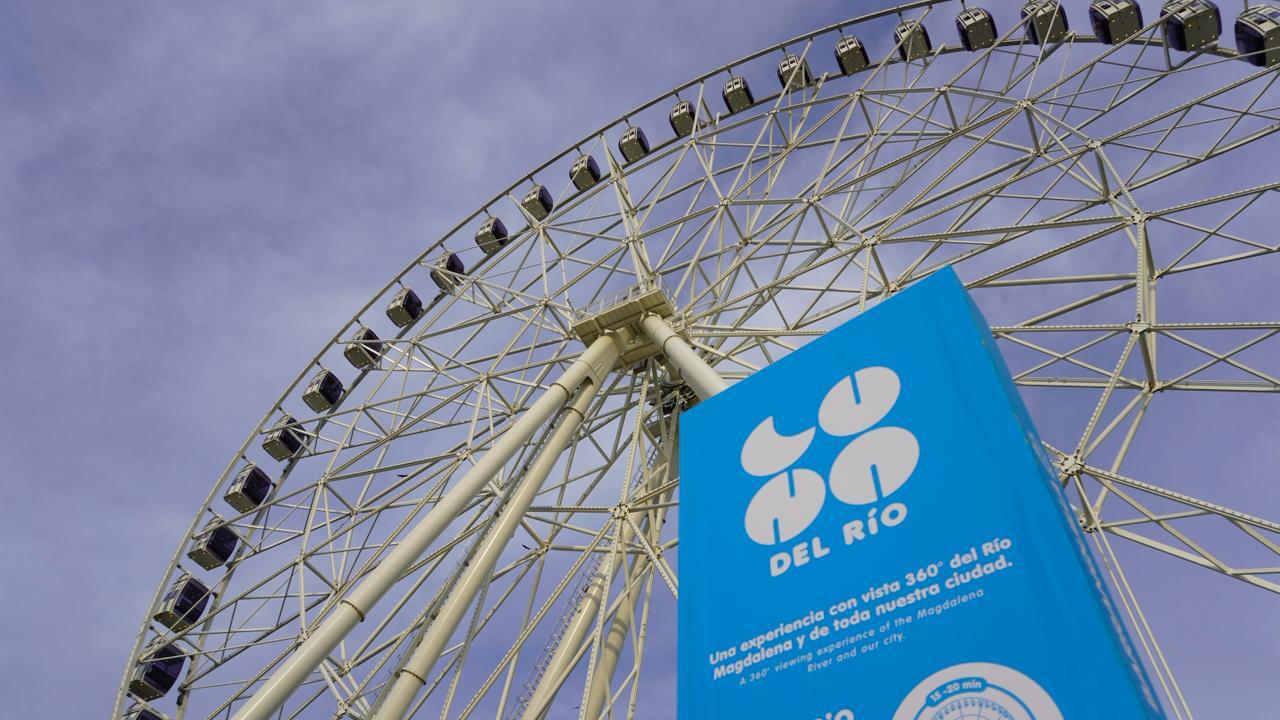 Una gran rueda de la fortuna metálica con cabinas distribuidas en su borde, vista desde un ángulo bajo contra un cielo despejado gris azulado. Hay un cartel azul con texto blanco que promociona la atracción, indicando que ofrece una experiencia con vista panorámica de 360° del Río Magdalena y la ciudad.