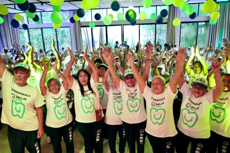 Un grupo de adultos mayores con camisetas blancas que dicen “Barranquilla Salud” y un logotipo verde levanta los brazos en un salón decorado con globos verdes y azules.