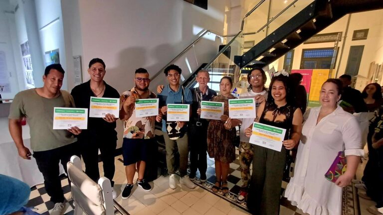 un grupo de personas reunidas en un espacio interior con diseño moderno y piso de baldosas en blanco y negro. Todas las personas están de pie y sostienen certificados impresos con encabezado verde y texto en negro.