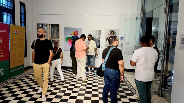 una exposición artística en un espacio interior con piso de baldosas en blanco y negro, paredes blancas y buena iluminación. En el centro y hacia el fondo, varias personas observan y comentan las obras exhibidas en la pared. Se distinguen al menos tres piezas de arte colgadas.