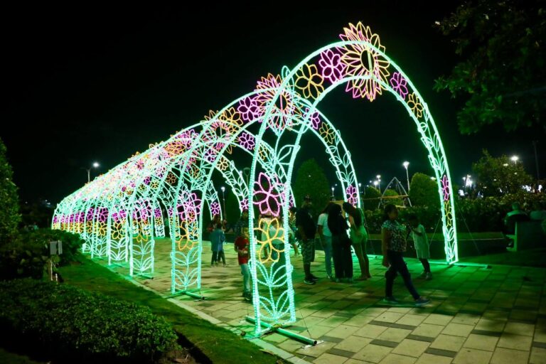 estructura decorativa iluminada en forma de arco, compuesta por múltiples secciones consecutivas que crean un túnel de luz. Cada arco está adornado con diseños florales en colores vivos.