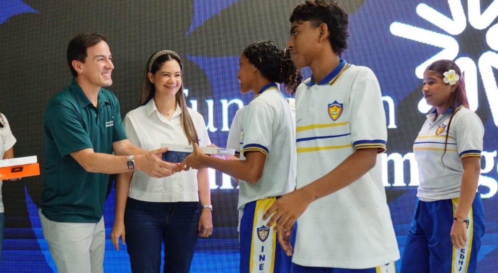 ceremonia de clausura en un escenario institucional con fondo azul y diseño gráfico. En primer plano, una persona entrega un reconocimiento a un estudiante que viste uniforme escolar blanco con detalles en azul y amarillo, acompañado de otros estudiantes con el mismo uniforme.