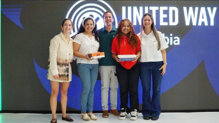 Cinco personas posando en un escenario institucional frente a una pantalla LED que exhibe el logo y texto “UNITED WAY Colombia” en letras blancas sobre un fondo azul oscuro . Dos personas sostienen cajas rectangulares, una de color naranja y otra azul, que son premios.