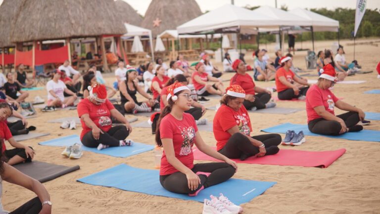 actividad grupal al aire libre en una playa, donde varias personas están sentadas sobre colchonetas de ejercicio, meditación o yoga. Todas las personas visten camisetas rojas con motivos navideños y llevan gorros de Navidad