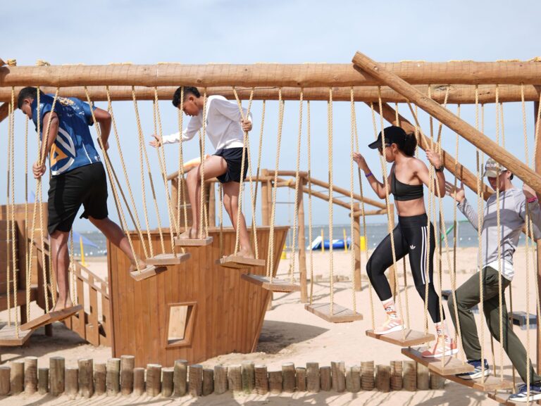 ctividad recreativa en la playa, donde cuatro personas están participando en un circuito de cuerdas y plataformas suspendidas. El escenario está compuesto por estructuras de madera con cuerdas que forman puentes colgantes, diseñados para pruebas de equilibrio y destreza física. 