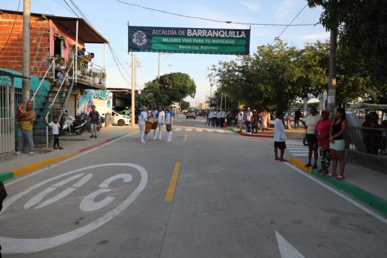  Calle recién pavimentada en un barrio residencial, decorada con señalización vial nueva, incluyendo una marca grande de límite de velocidad pintada sobre el asfalto. A ambos lados de la vía, varias personas se encuentran reunidas.