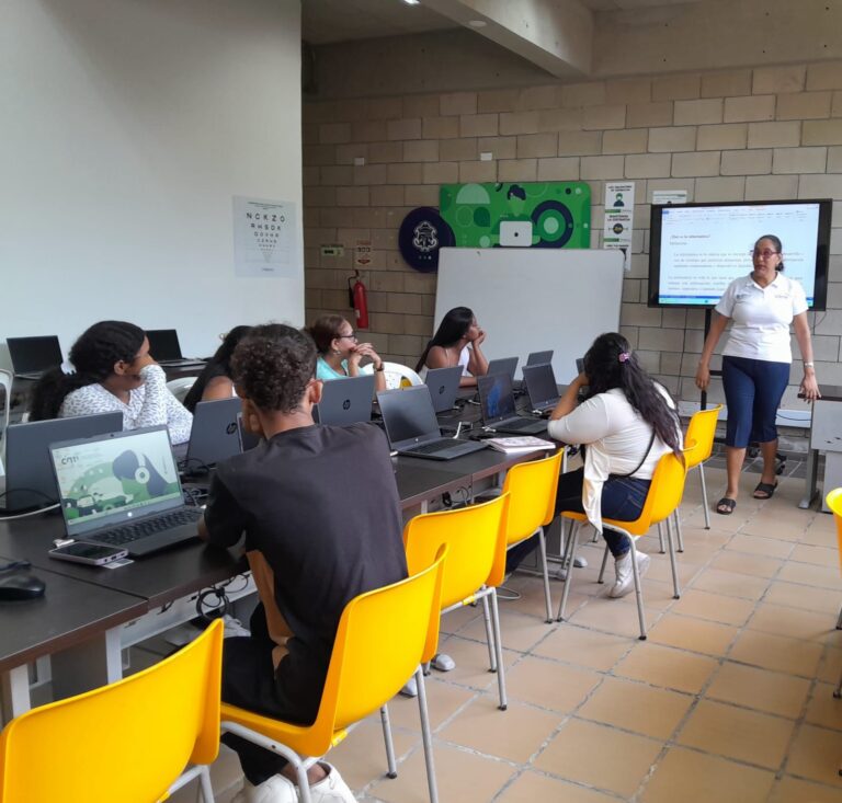 Sala de informática con varias mesas largas equipadas con computadoras portátiles. Al frente, una pantalla de gran formato proyecta contenido digital mientras la profesora explica, y los estudiantes observan con atención
