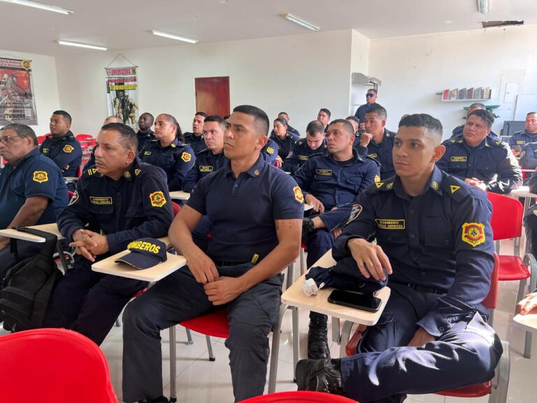  Bomberos sentados en filas, en sillas rojas con mesas incorporadas, usando su uniforme institucional. El espacio es amplio y bien iluminado, y en las paredes hay afiches sobre prevención y seguridad. Se trata de una jornada de capacitación o reunión para el personal del cuerpo de bomberos.
