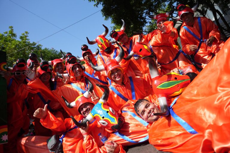 Un grupo de comparsa Todas llevan trajes de color naranja brillante con cintas azules cruzadas en el pecho y la cintura. Además, cada persona sostiene o porta una máscara en forma de toro, muy colorida y llamativa.