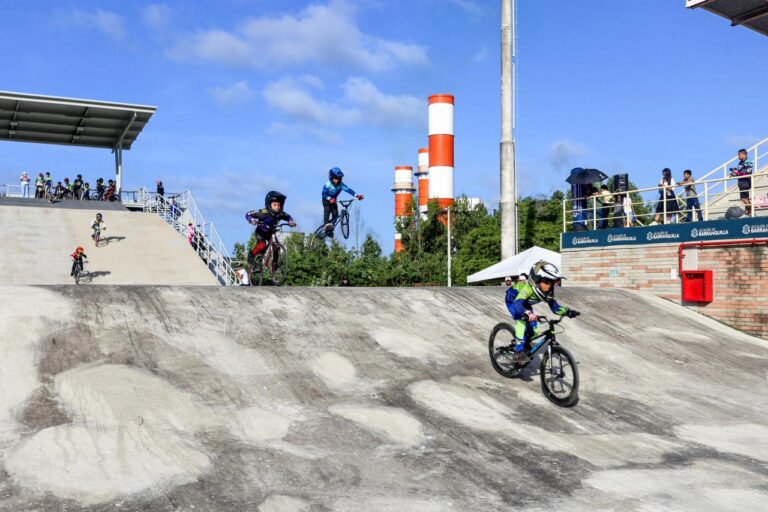 Ciclista con uniforme verde y azul desciende por la rampa. En la parte superior, otros ciclistas están realizando saltos en el aire con sus bicicletas, usando cascos y equipo de protección.