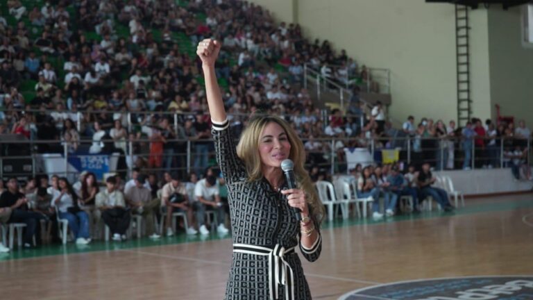 Ana Maria Aljura con un micrófono on una mano y levantando el puño con la otra en señal de ánimo y celebración, mientras se dirige a un público numeroso que llena las graderías.
