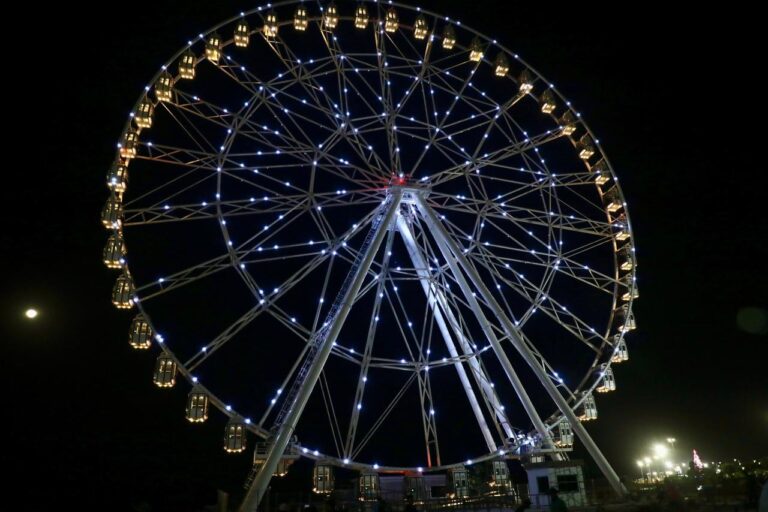 gran rueda de la fortuna iluminada durante la noche. Su estructura metálica está detalladamente resaltada por luces blancas y cálidas distribuidas a lo largo de los radios y las cabinas. En la parte inferior se observan las bases que sostienen la rueda, también iluminadas. 