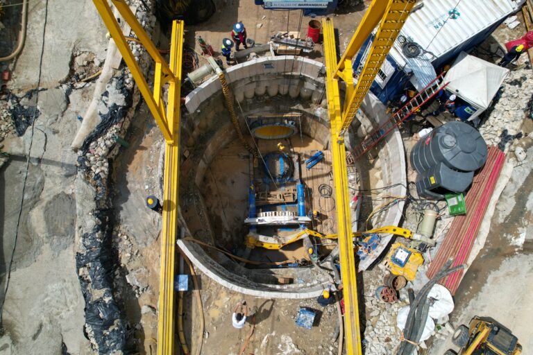Obra de infraestructura vista desde arriba, específicamente un pozo circular profundo en construcción. El pozo está rodeado por un muro de concreto segmentado y dentro se observan equipos pesados de color azul, posiblemente maquinaria para perforación