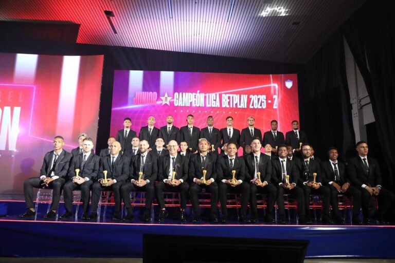 Grupo de futbolista y técnico del equipo de futbol, vestidas de manera formal, con trajes oscuros y corbatas, sentadas y de pie en un escenario iluminado, sosteniendo un reconocimiento