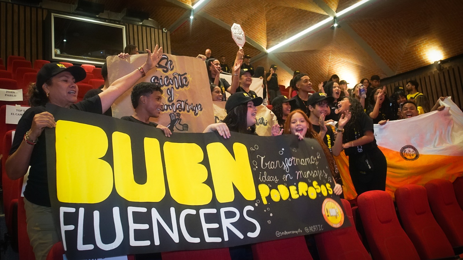 Jóvenes y docentes portando alegremente un cartel de Buenfluencer en un auditorio