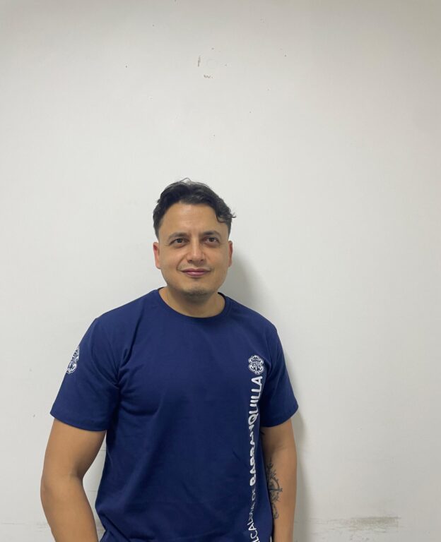 Funcionario con vestimenta institucional, de pie en plano medio con pared blanca de fondo