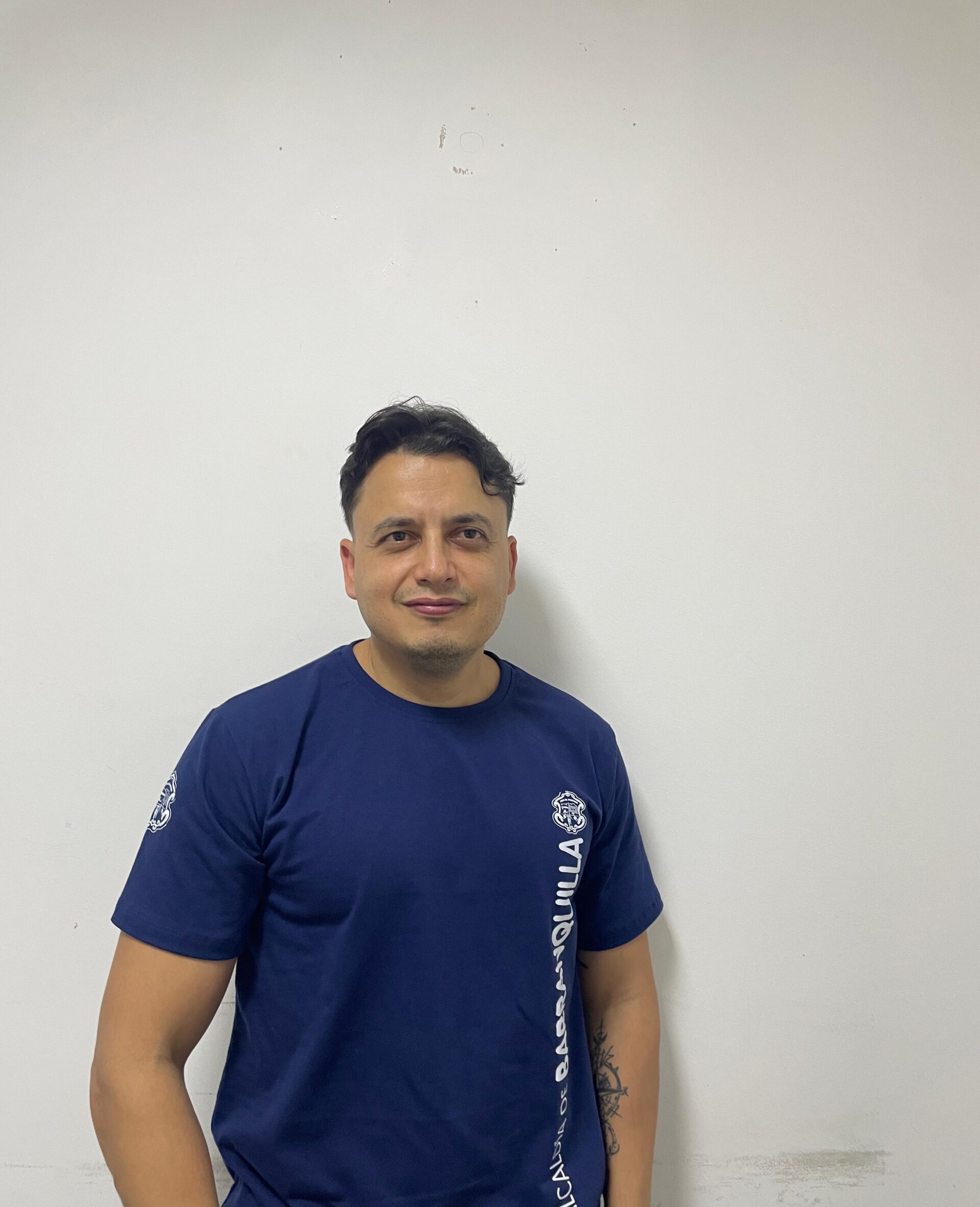 Funcionario con vestimenta institucional, de pie en plano medio con pared blanca de fondo